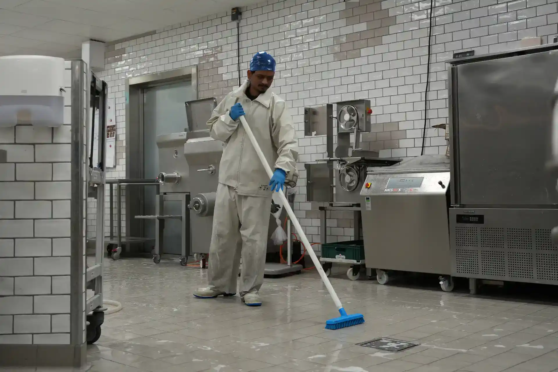 Person rengør gulv i fødevarefabrik med moppe og beskyttelsesudstyr.