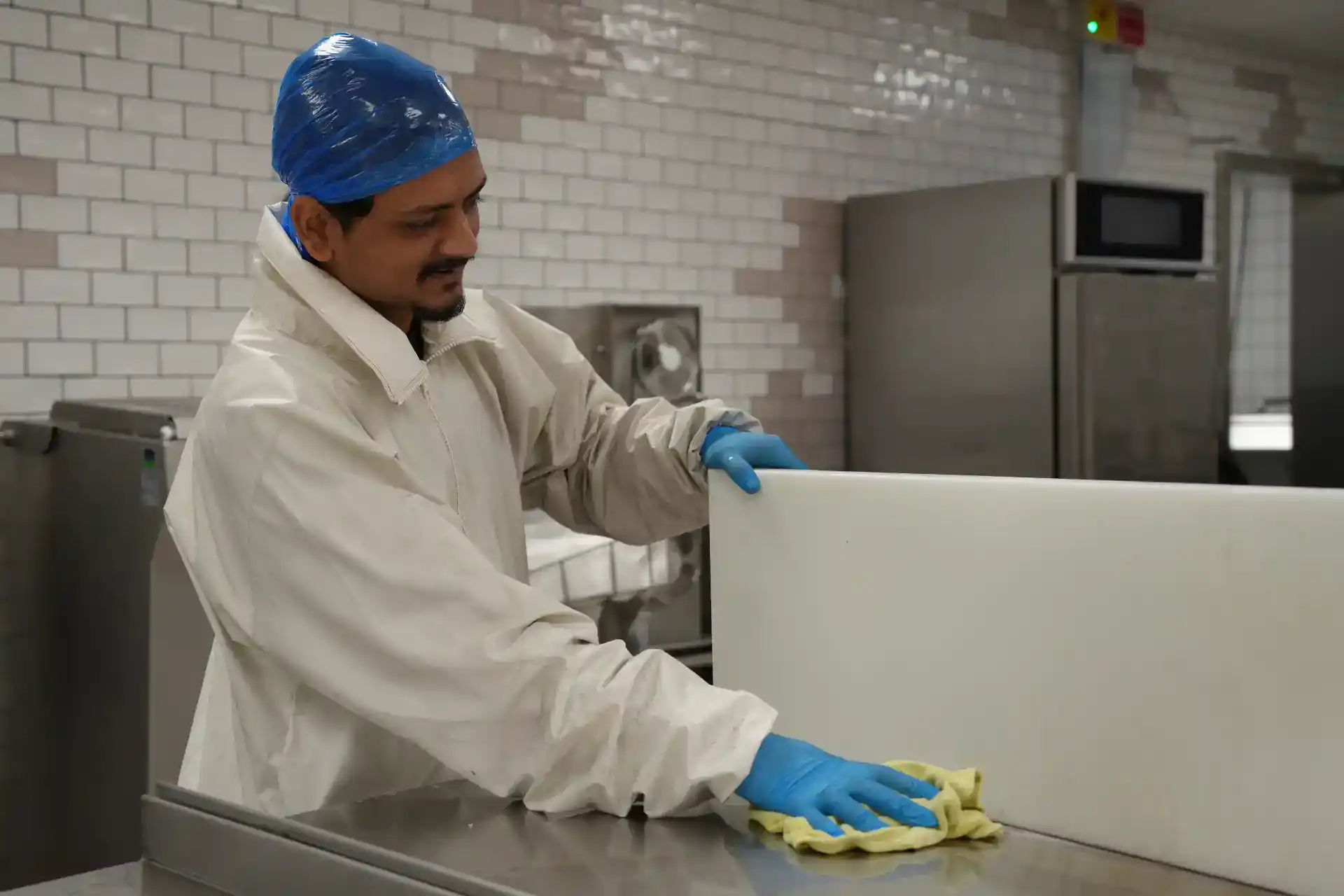 Mand i rengøringsdragt rengør industrikøkken med klud og rengøringsmiddel.