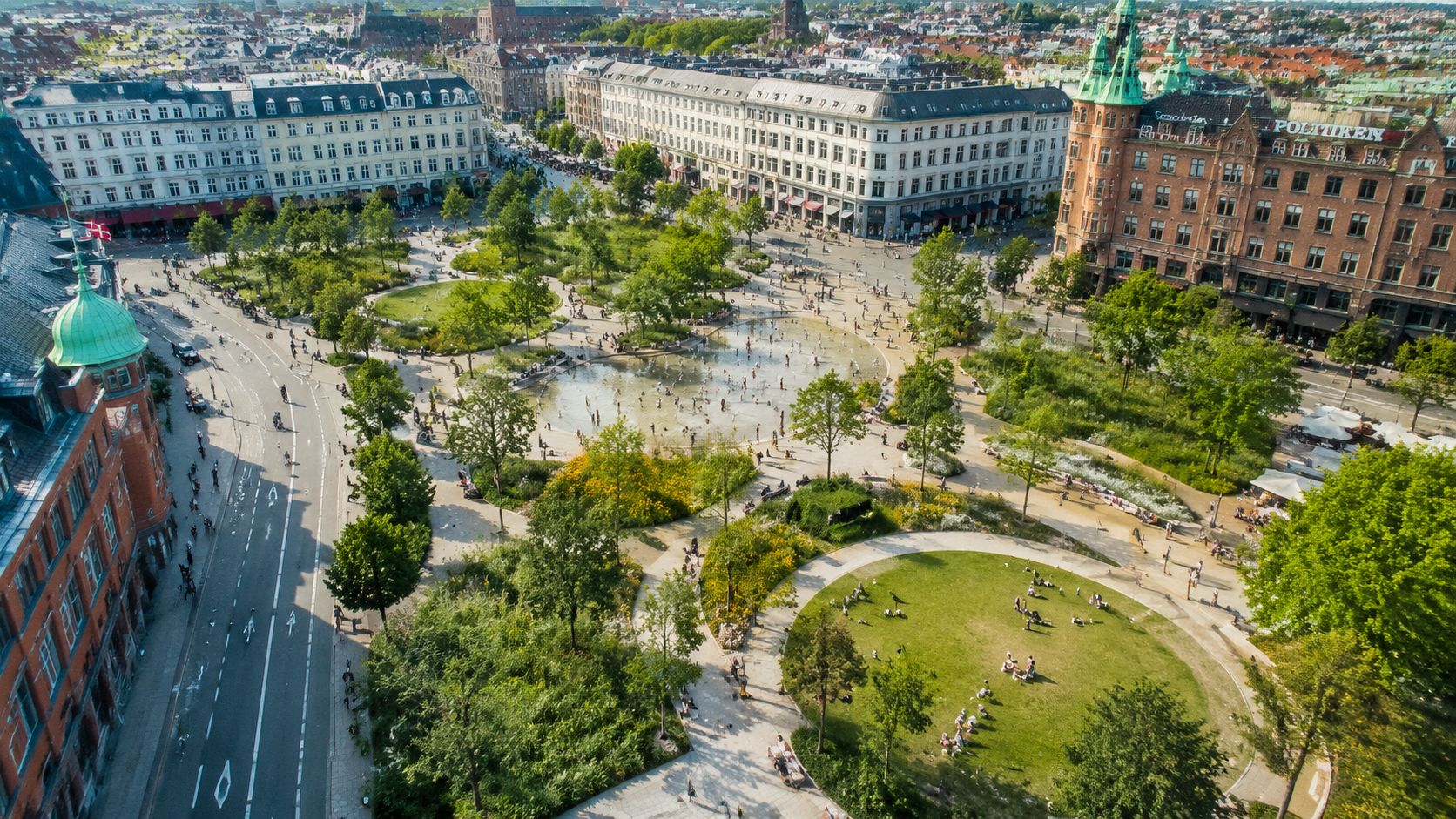 Set oppefra af Israels Plads i København med grønne områder, vandfontæner, legende mennesker og historiske bygninger i baggrunden.